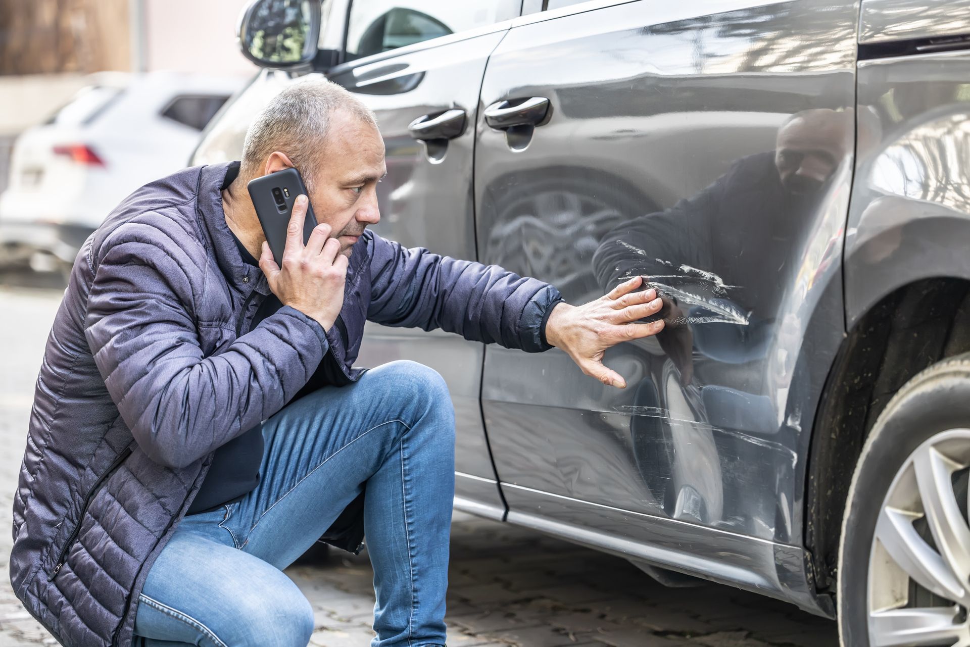 A man on the phone kneels next to a car, examining and touching a noticeable scratch on the vehicle’s door—likely seeking advice from Illinois Insurance Center about his coverage options.