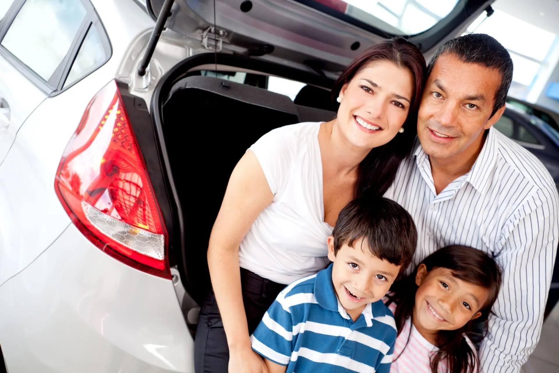 A smiling family of four poses together by the open trunk of a silver car. Thanks to Illinois Insurance Center, the parents and their two children— a boy in a striped shirt and a girl in pink—enjoy peace of mind, all looking happily at the camera.