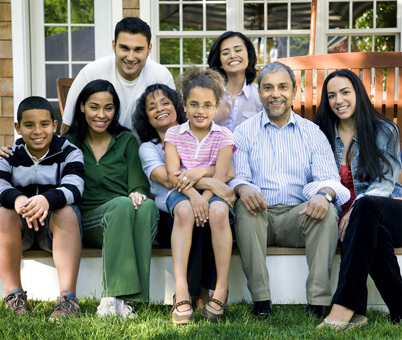 A multigenerational family of eight, including adults and children, sits and smiles together outside on a bench and steps in front of a house, enjoying a sunny day and peace of mind with Illinois Insurance Center.