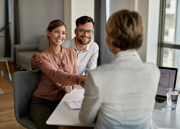 A smiling couple sits across a desk from a professional woman at Illinois Insurance Center, shaking hands. Papers and a laptop on the desk suggest a successful business or financial meeting in a bright, modern office.