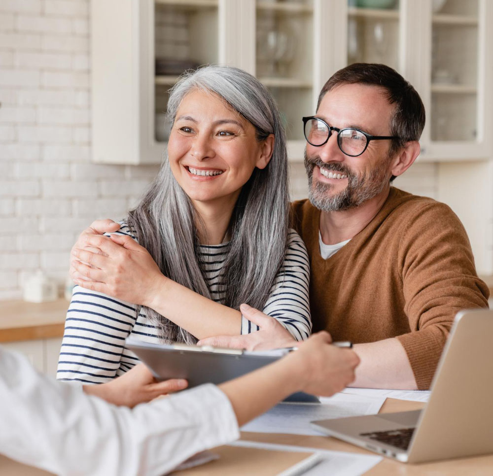 A smiling middle-aged couple sits together at a table, embracing and listening to a person from the Illinois Insurance Center holding a clipboard. Papers and a laptop on the table suggest a friendly consultation in a bright kitchen.