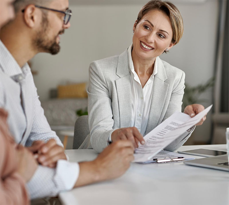 A smiling woman in a light-colored blazer holds and discusses a document with two people sitting across from her at a desk in the bright Illinois Insurance Center office.