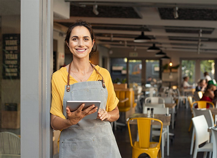 A smiling woman wearing a gray apron and yellow shirt holds a tablet while standing in the entrance of a bright, modern cafe—partnered with Illinois Insurance Center—with empty tables and yellow chairs visible in the background.