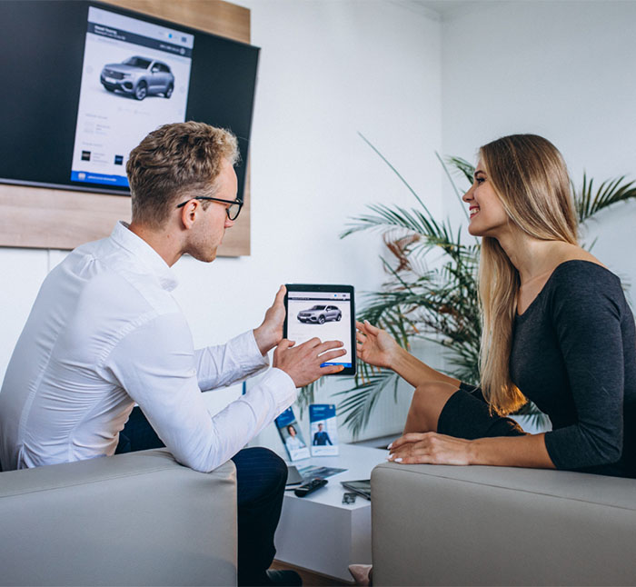A man and woman sit in a modern office at the Illinois Insurance Center, looking at a tablet displaying a car, with a matching car image shown on a large screen in the background. The woman is smiling and pointing at the tablet.