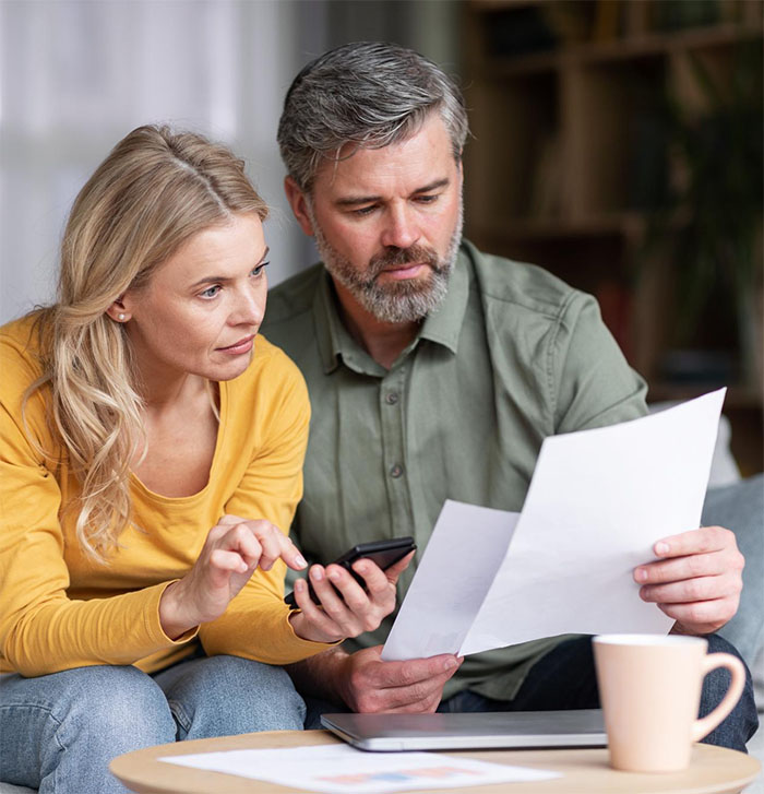A man and woman sit together on a couch, looking at documents from the Illinois Insurance Center. The woman holds a phone and points at the papers while the man looks focused. A coffee mug sits on the table in front of them.