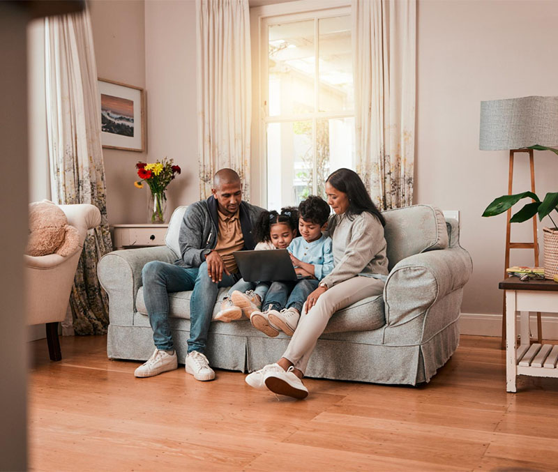 A family of four sits together on a light gray couch in a cozy living room, smiling and looking at a laptop. Sunlight streams through the window behind them as they explore coverage options from Illinois Insurance Center. Flowers decorate a table nearby.