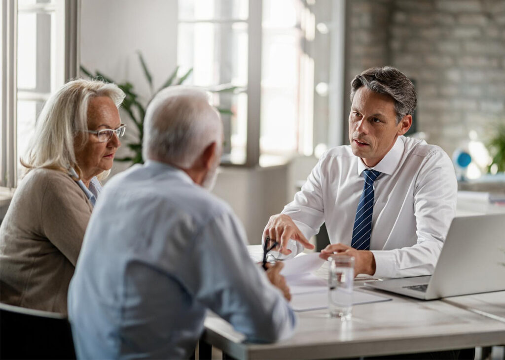 A businessman in a white shirt and tie sits at a desk, talking to an older couple at the Illinois Insurance Center. The couple listens intently while he gestures toward some documents, with a laptop and glass of water on the table.