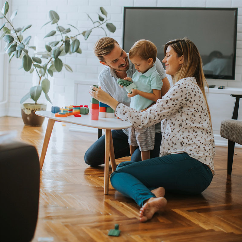 A smiling family of three sits on the floor in their living room, playing with colorful building blocks at a small table. The Illinois Insurance Center helps protect joyful moments like these for families just like yours.