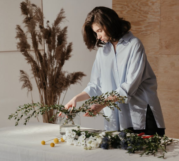 A person arranging green branches and flowers in a glass vase on a table, with various flowers and tools nearby—much like someone carefully selecting insurance in Burr Ridge. Tall dried plants stand in the background against a light wooden wall.