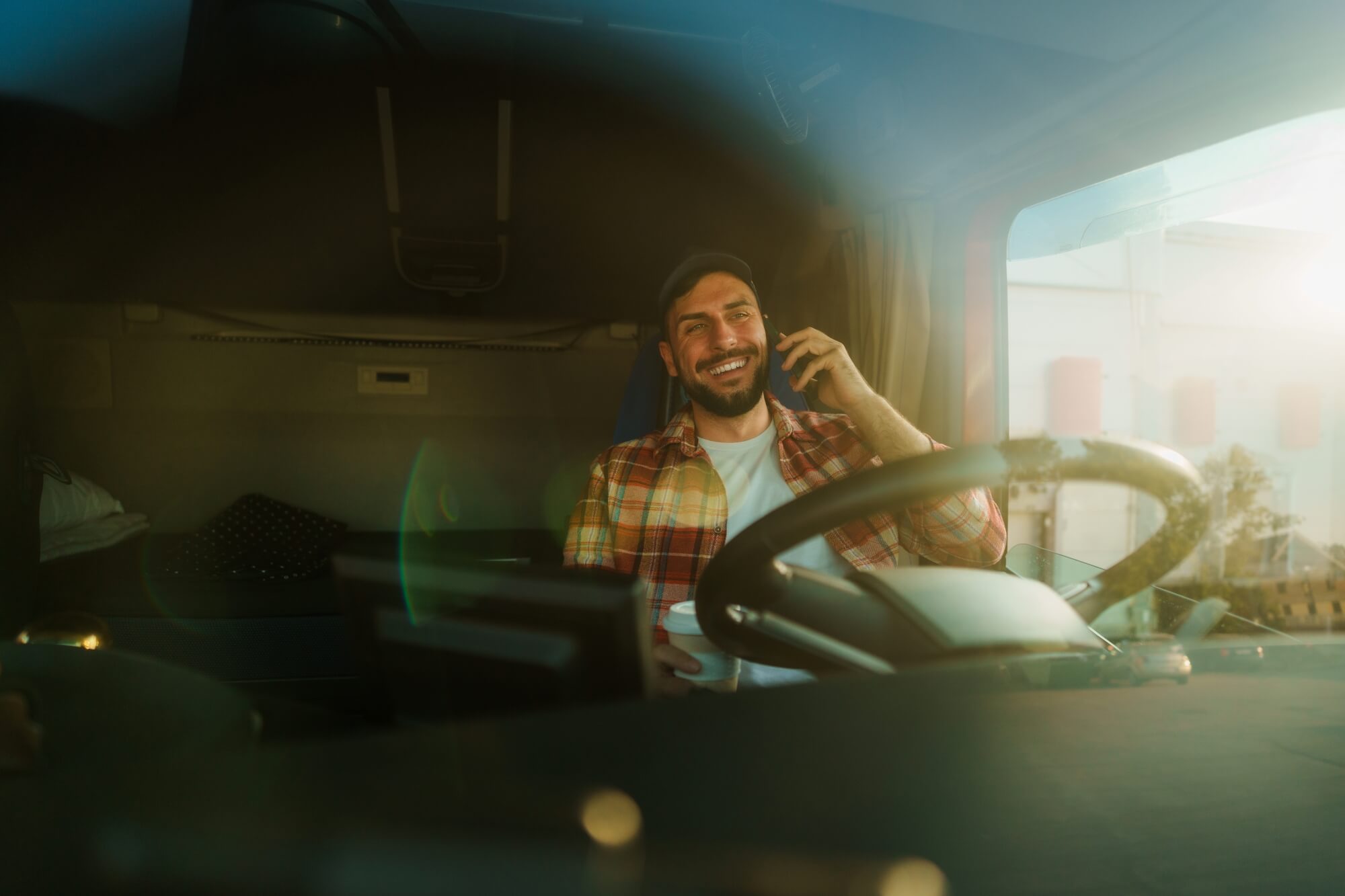 A man sitting in a truck cabin smiles while talking on his smartphone. Sunlight streams through the window, highlighting his relaxed, cheerful expression. The steering wheel and dashboard are visible, reminding him of the value of Commercial Truck Insurance.