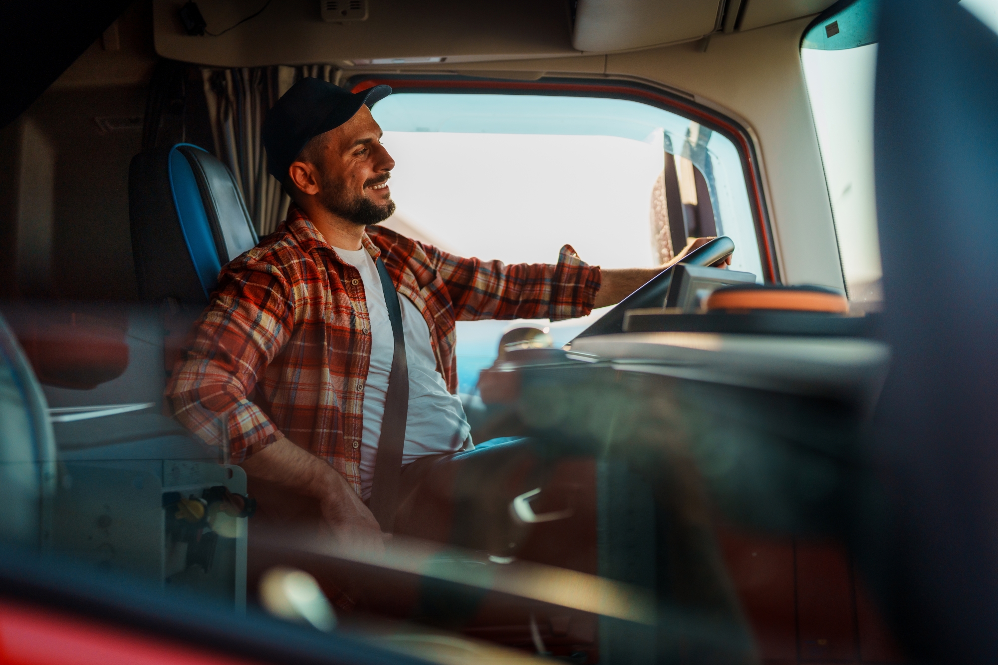 A smiling man wearing a cap and plaid shirt sits in the driver’s seat of a truck, holding the steering wheel and looking out the window, with sunlight streaming into the cab—ready for another day with Commercial Truck Insurance in Illinois​.