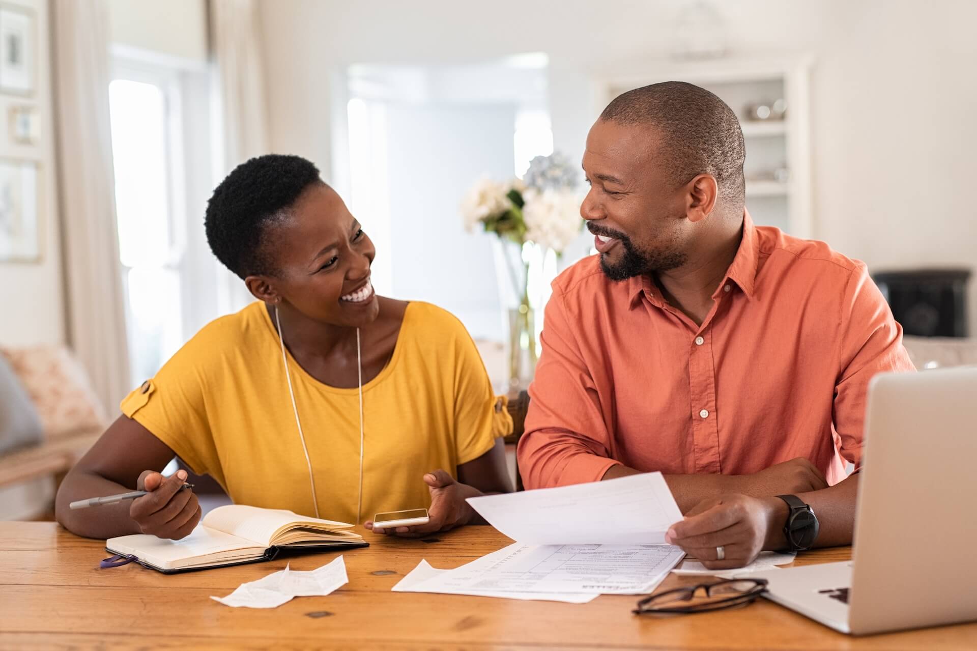 A smiling couple sits at a table with papers, a notebook, and a laptop, appearing to discuss finances or planning together in their bright, cozy home—perhaps after seeing an Exit Intent Popup with helpful budgeting tips.