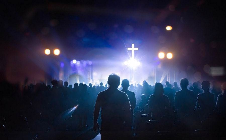 A crowd of people stands in a dark room facing a brightly lit stage with a glowing white cross, as blue and purple lights illuminate the scene—highlighting the importance of Church Insurance for such vibrant gatherings.
