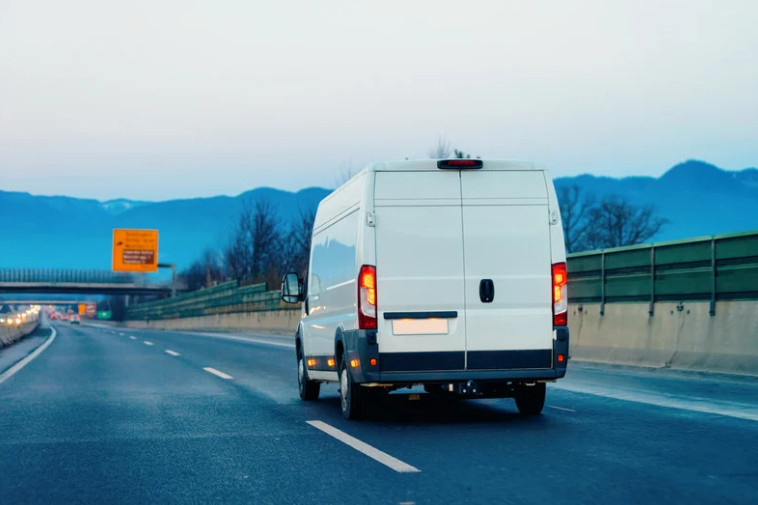 A white delivery van, protected by Commercial Auto Insurance, drives on an empty highway with mountains in the background and a road sign to the left. The clear sky has a slight blue tint.