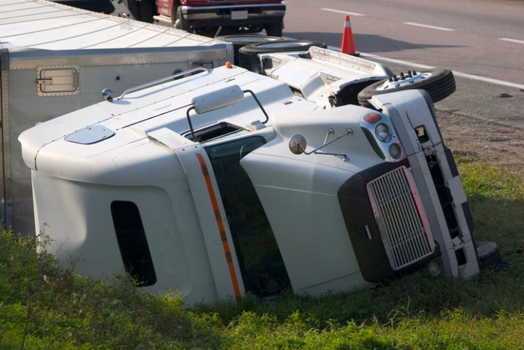 A white semi-truck is overturned on its side off the road beside a highway, with traffic cones and other vehicles visible in the background—a scene highlighting the importance of commercial truck insurance.