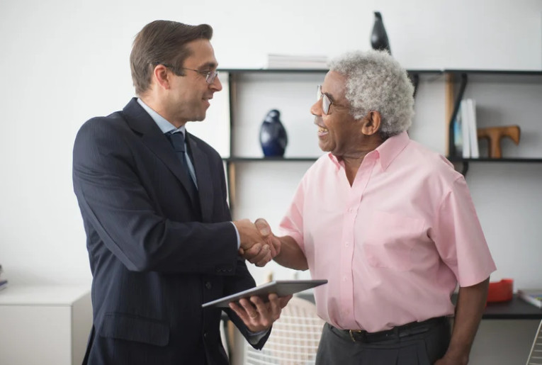 Two men, one in a suit holding a tablet and the other in a pink shirt, smiling and shaking hands in an office setting—celebrating their new renters insurance policy amid shelves and decorative items.