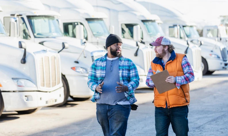 Two men, one holding a clipboard, walk and talk in front of a row of white semi-trucks parked outdoors. Both are dressed in casual, warm clothing and appear to be discussing commercial truck insurance for their fleet.