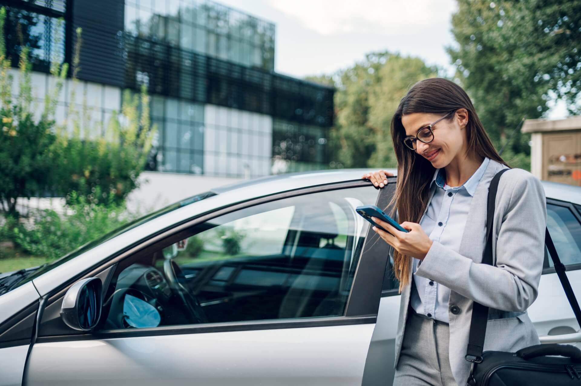 A woman in business attire stands by an open car door, smiling while looking at her smartphone—perhaps checking her auto insurance Rockford IL policy—with a modern glass building and greenery in the background.