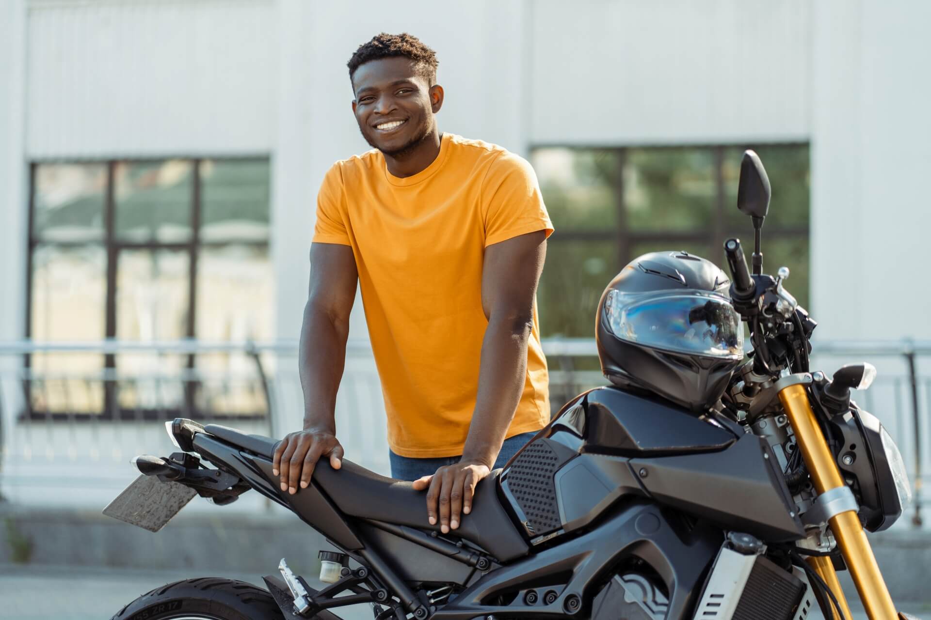 A man in a bright orange t-shirt smiles while standing outdoors with his hands resting on the seat of a black motorcycle, showcasing his ride. If you need auto insurance Schaumburg IL, make sure your bike is protected on every adventure.