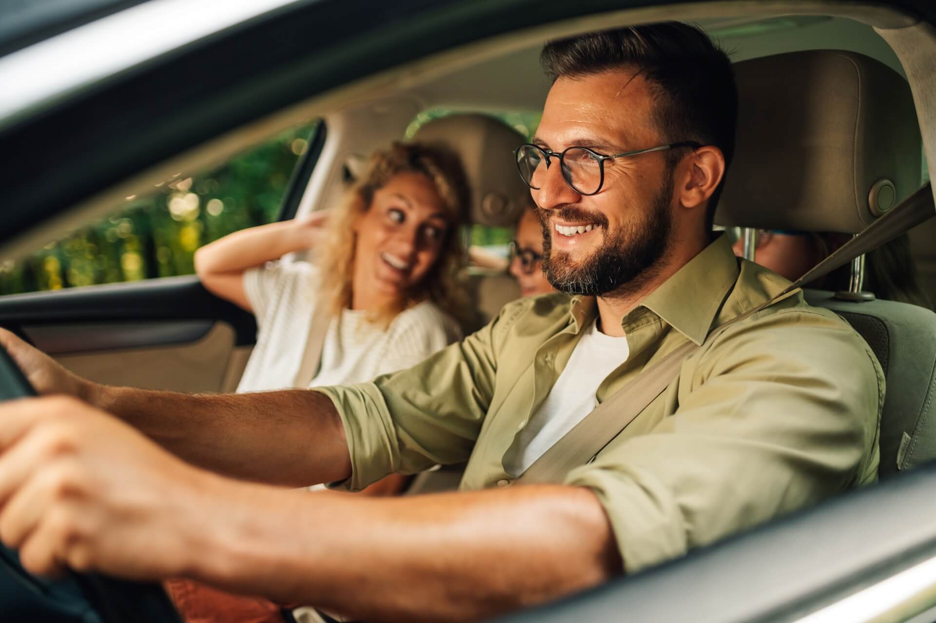 A smiling man drives a car with a woman and a child in the back seat. The woman is laughing and looking at him, while sunlight streams through the window—perfect moments protected by auto insurance Elgin IL.
