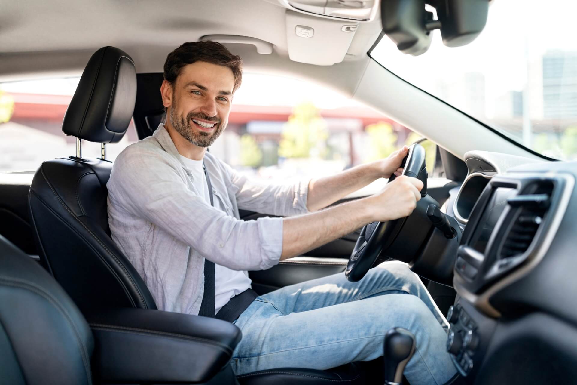 A smiling man with a beard sits in the driver’s seat of a car, holding the steering wheel with both hands. He is wearing a light shirt and jeans, enjoying the sunlight—safe and secure with auto insurance Elgin IL.