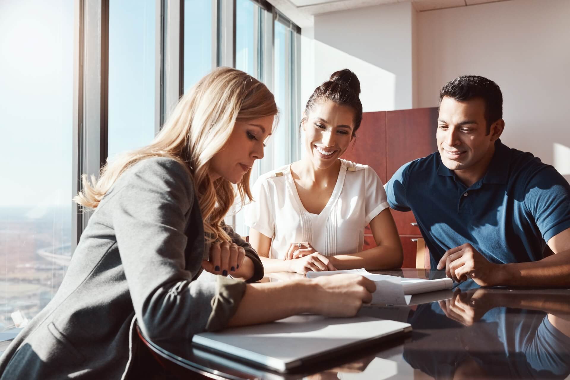 Three people sit at a glass table in a sunlit office, discussing auto insurance Rockford IL. A woman in a blazer writes while a smiling woman and man sit across from her, appearing engaged and attentive.