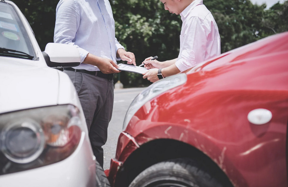 Two people stand between a white car and a red car that appear to have collided. They are holding a clipboard and pen, likely exchanging information or filing a car insurance report after the accident.