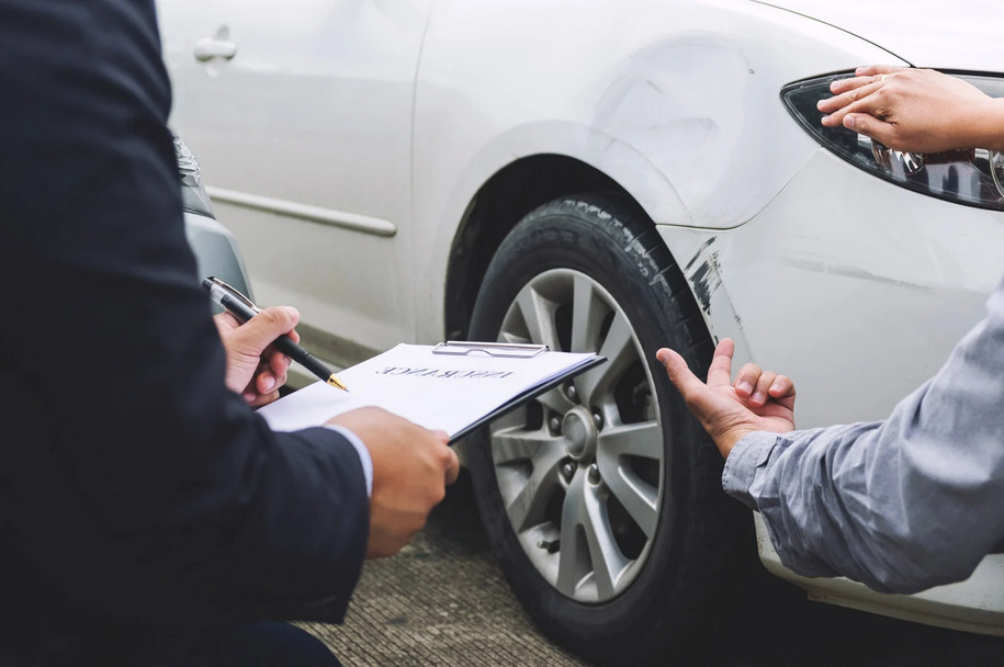 Two people inspect the scratched front fender of a white car; one holds a clipboard with paperwork, likely assessing the damage for an Illinois car insurance claim.