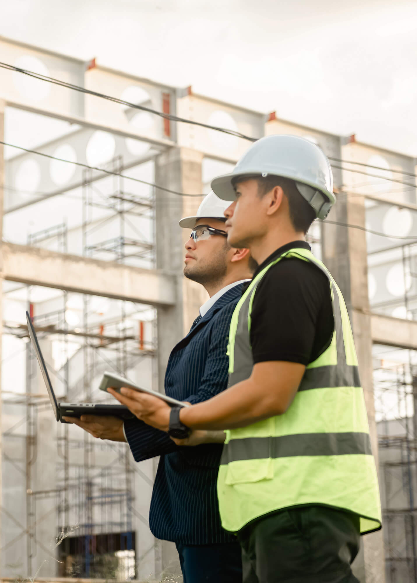 Two men wearing safety helmets stand outdoors at a construction site; one in a safety vest holds a laptop, while the other, dressed in a suit, discusses building framework—highlighting the importance of builders risk insurance Illinois.