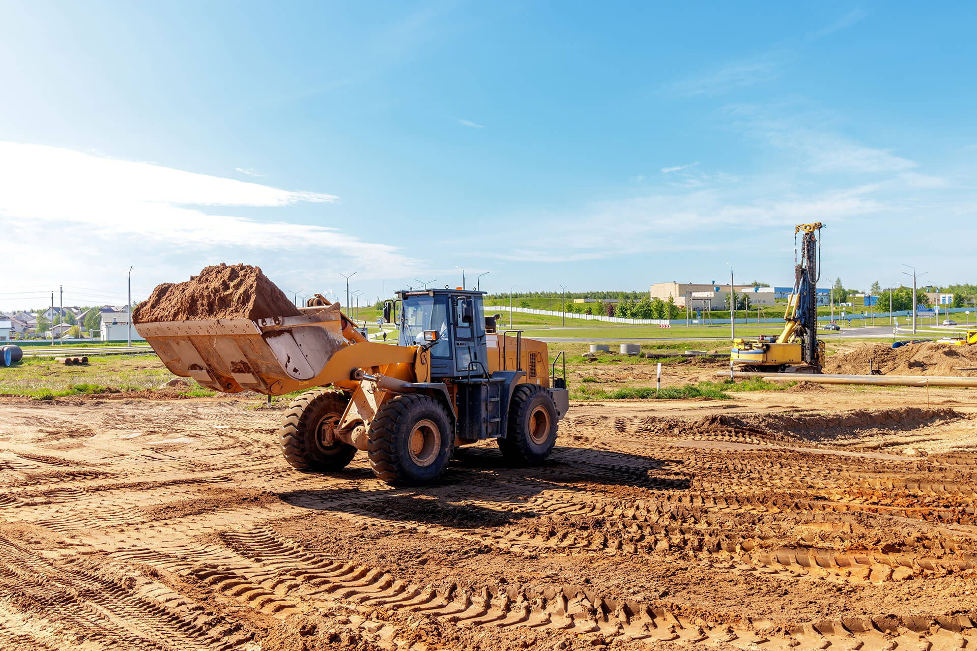 A large yellow front loader carries a load of dirt at a construction site with visible tire tracks, while a drilling machine and buildings stand in the background under a clear blue sky—ideal for those considering builders risk insurance Illinois.