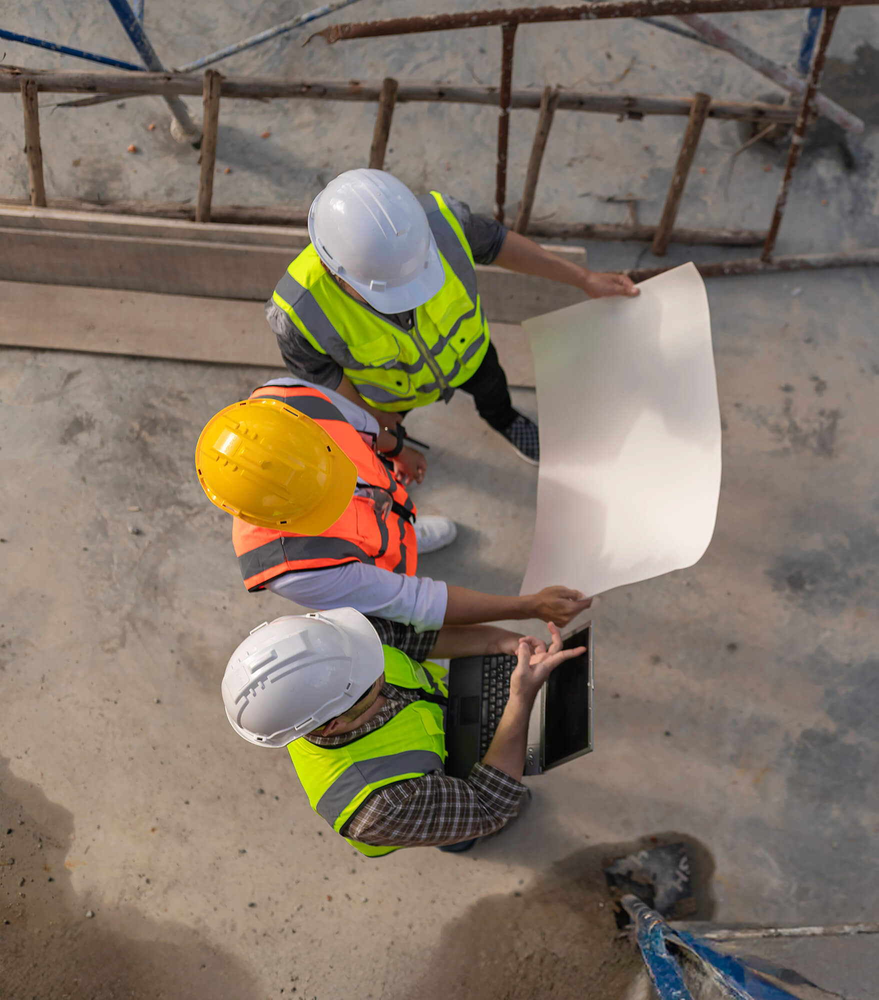 Three construction workers in safety vests and helmets stand on a site in Illinois, reviewing a large blueprint and laptop. The image is taken from above, highlighting the importance of builders risk insurance Illinois on the unfinished concrete ground.