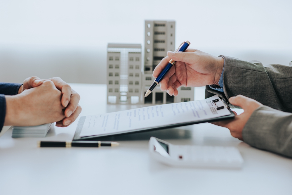 Two people sit at a desk, one holding a clipboard with a form and pen, the other with hands clasped; model buildings and a calculator highlight their discussion about Commercial Property Insurance in Illinois.