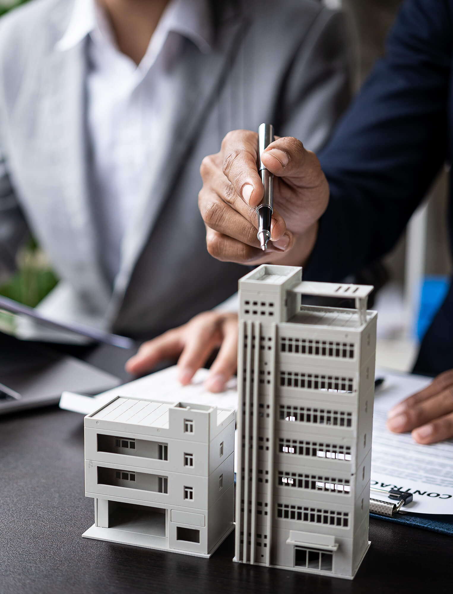 Two people in business attire sit at a desk with documents, one holding a pen and gesturing towards two architectural building models, as they discuss Commercial Property Insurance Illinois.