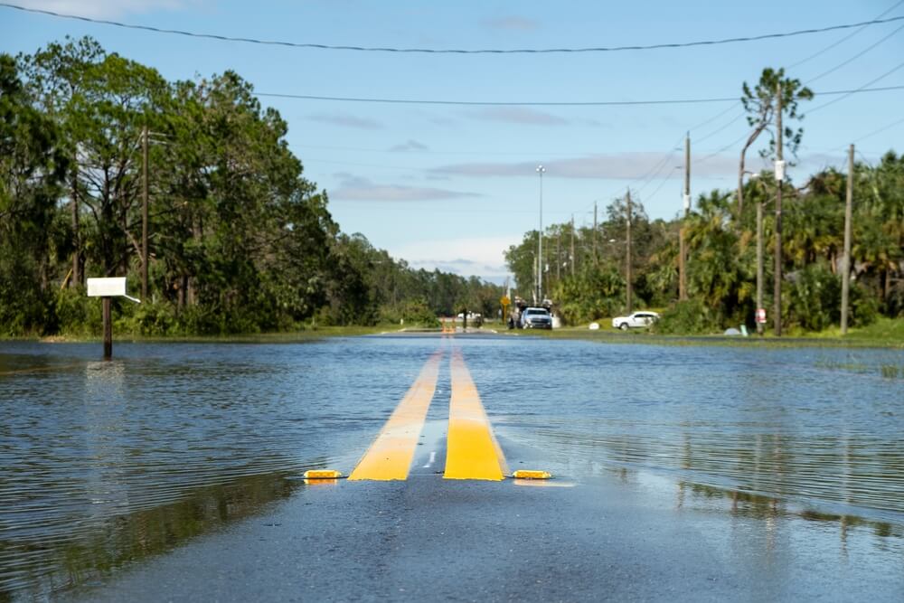 A flooded road with double yellow lines is partially submerged under water, surrounded by trees, utility poles, and a mailbox—a reminder of why Addison homeowners insurance can be essential. A few vehicles and people are visible in the background.