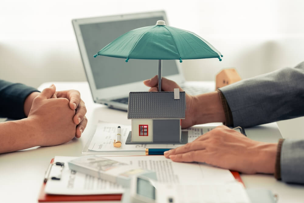 Two people sit at a desk with documents, a calculator, and a laptop. One person holds a small house model under a green umbrella, symbolizing Addison homeowners insurance and the importance of reliable home protection.