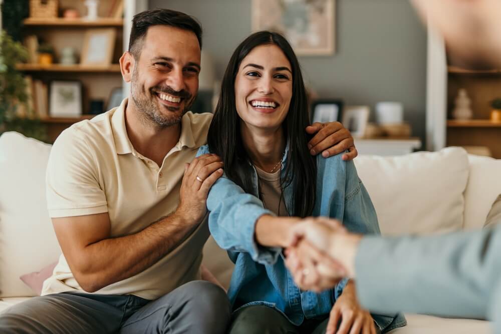 A man and woman sit together on a couch, smiling and making eye contact as they shake hands with someone—perhaps discussing Addison homeowners insurance—in a cozy space with books and decor on the shelves behind them.