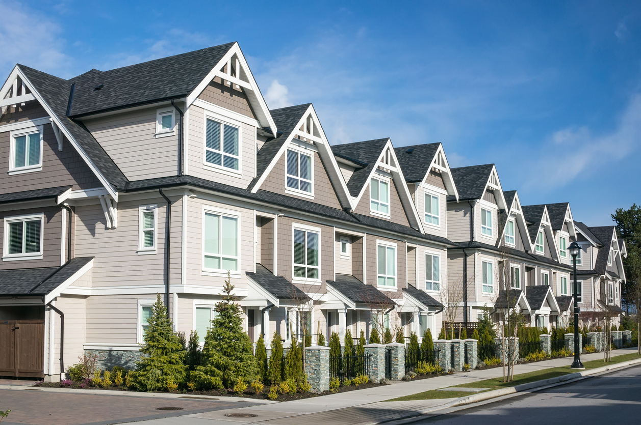 A row of modern, multi-story townhouses with gabled roofs and large windows lines a clean street in Alton IL, where small shrubs and trees add charm. Protect your investment with homeowners insurance under the clear blue sky.