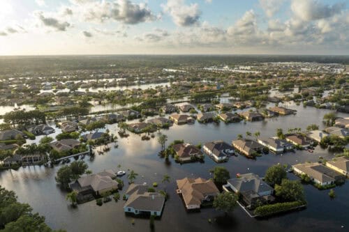 Aerial view of a suburban neighborhood in Antioch, IL, with many homes partially submerged in floodwaters—highlighting the importance of homeowners insurance—surrounded by trees and sunlight beneath a partly cloudy sky.