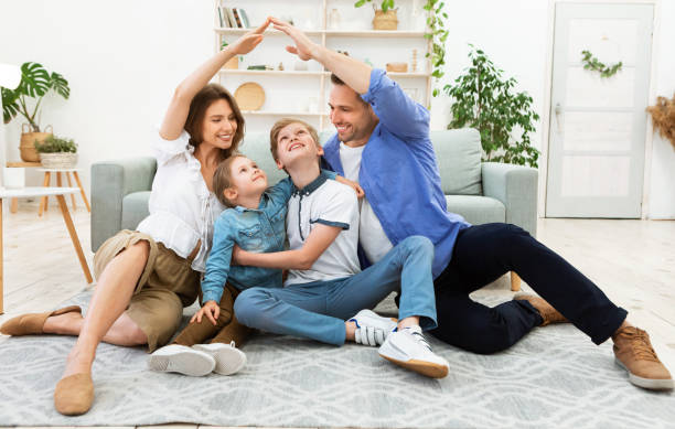 A family of four sits on a rug in a bright Alton IL living room. The parents form an arch with their arms above the two children, who are smiling and looking up. A sofa, shelves, and plants are visible in the background, highlighting happy homeowners.