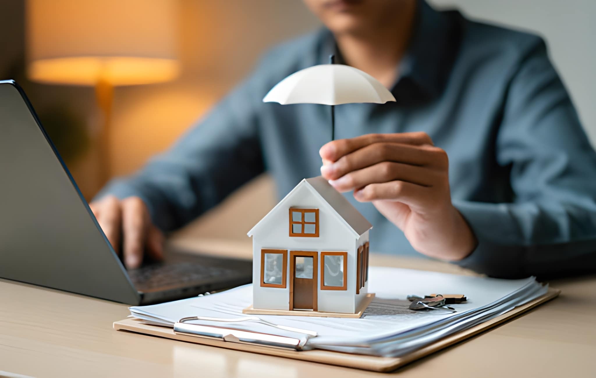 A person holds a small white umbrella over a model house placed on documents with a clipboard and keys, highlighting the importance of algonquin homeowners insurance while working on a laptop at a desk.