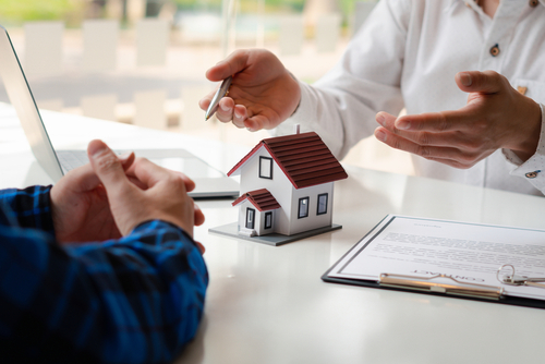 Two people sit at a table in Alsip, IL, discussing real estate and homeowners insurance, with a small model house, a contract, and a pen on the table. One gestures with the pen while the other listens attentively with hands clasped.