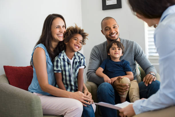 A smiling family of four sits on a couch during a meeting about Homeowners Insurance in Antioch IL. The parents and two young children face the professional holding papers, who is partially visible in the foreground.
