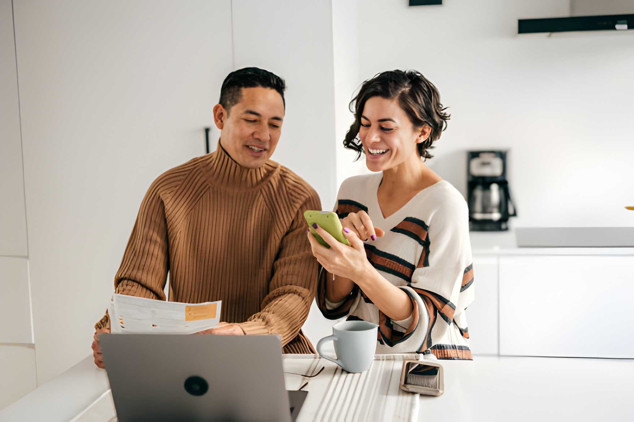 A man and a woman sit at a kitchen counter. The man holds a document, possibly about Algonquin homeowners insurance, while the woman smiles using her smartphone. A laptop, mug, and phone rest on the counter in front of them.