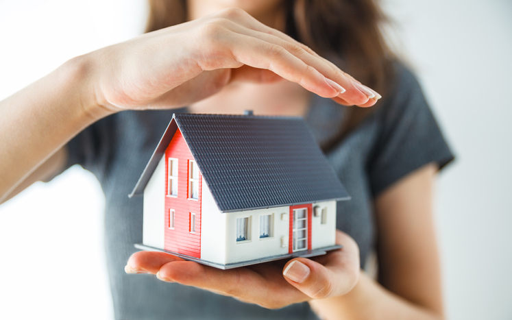 A person holds a small model house with both hands, one supporting the base and the other protectively over the roof—symbolizing the security homeowners insurance offers in Antioch IL. The model house is white with a red accent and a dark roof.