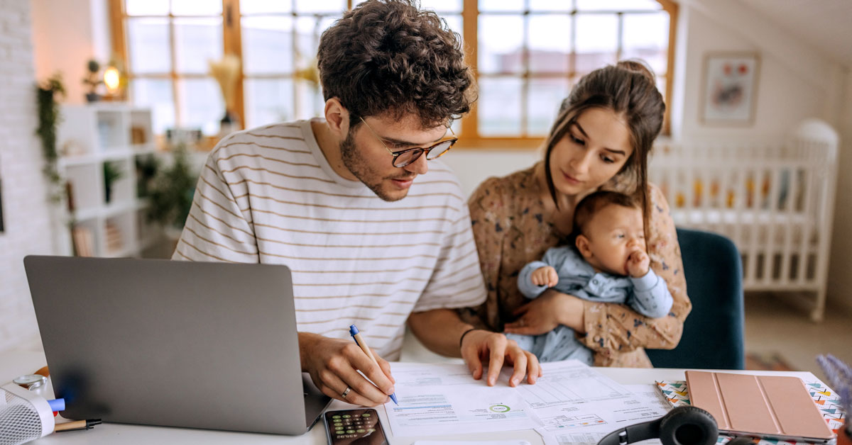 A man reviews documents at a desk with a laptop and calculator while a woman holding a baby looks on. The scene takes place in a bright Bensenville IL home office with large windows, a crib, and homeowners insurance papers in the background.
