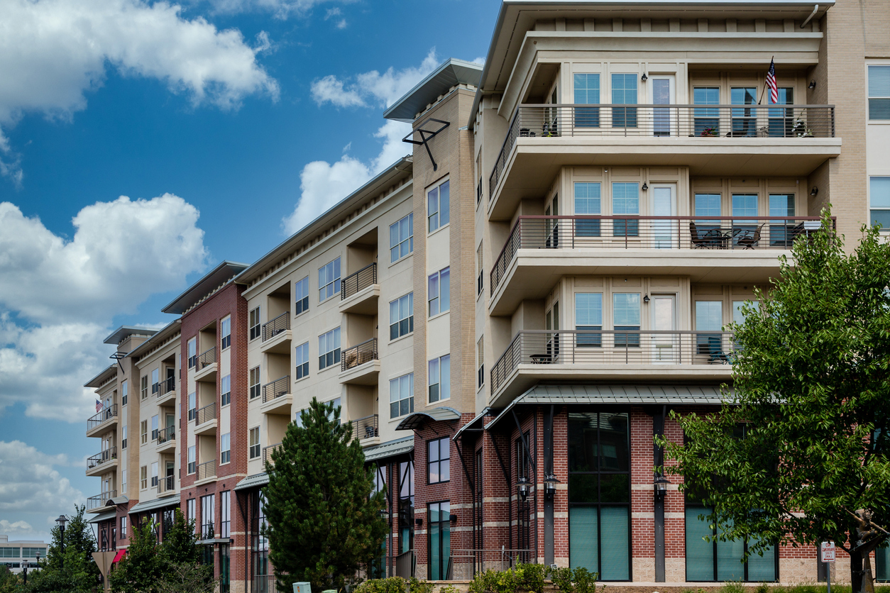 A modern multi-story apartment building with balconies, large windows, and a blend of brick and light-colored siding in Beach Park IL. Trees and bushes fill the foreground under a partly cloudy sky—ideal for those seeking homeowners insurance.