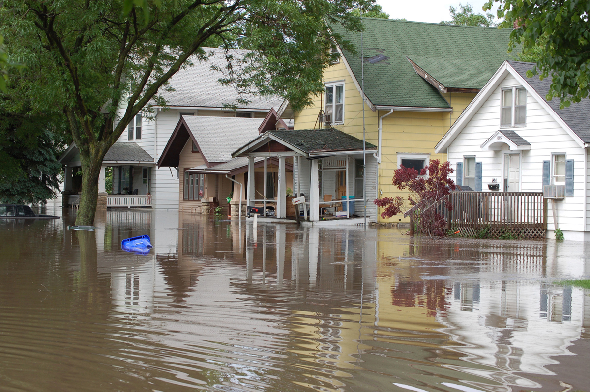 Floodwater surrounds several houses in this Beach Park IL neighborhood, submerging streets and reaching porches. A large tree and a tipped blue plastic container float nearby, highlighting the need for reliable homeowners insurance.