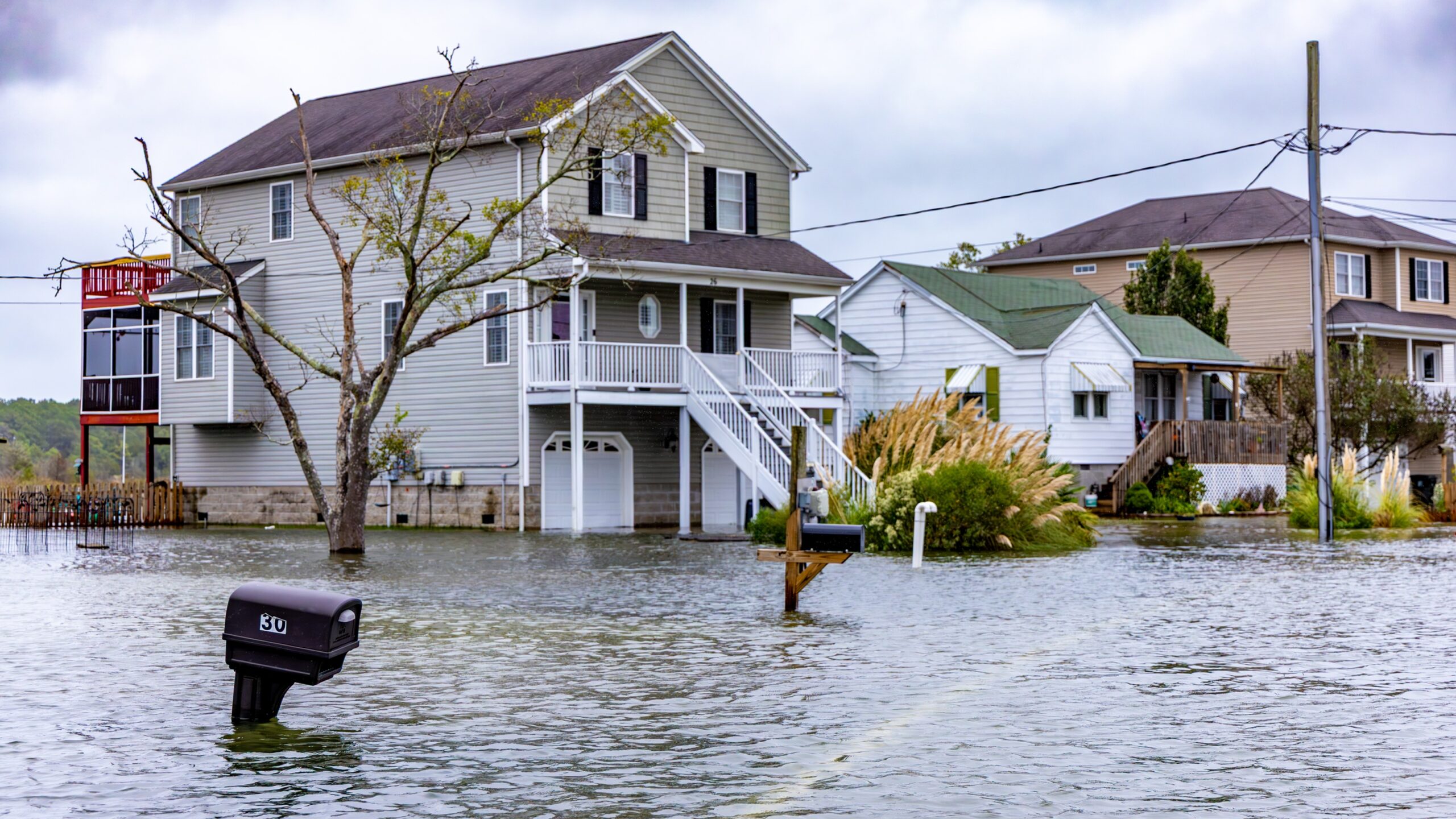 Flooded residential street in Belleville IL with several houses partially submerged in water. Mailboxes and a tree are surrounded by floodwater, highlighting the importance of having homeowners insurance. Cloudy skies appear overhead.