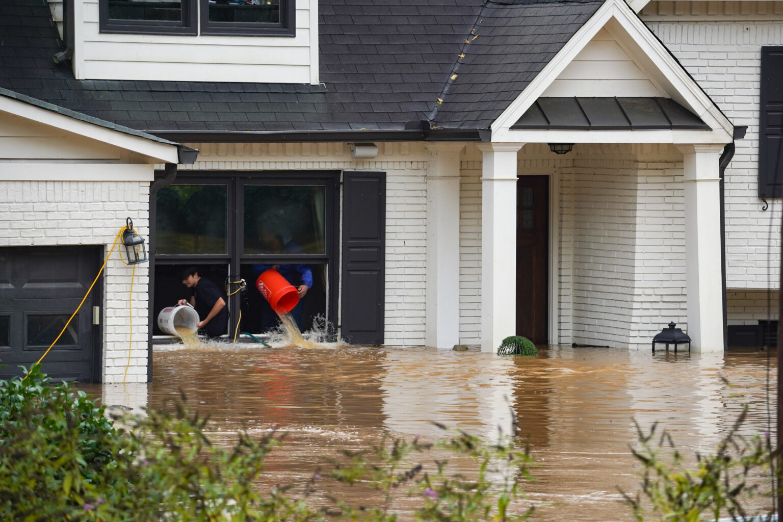 Two people in a flooded white house with dark trim in Bellwood, IL use buckets to bail out water through a window. Homeowners insurance could help protect against damages from high floodwaters covering the yard and reaching the windows.