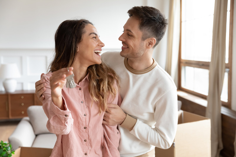A smiling woman holding house keys stands next to a man in a bright Bellwood, IL living room with unpacked boxes in the background. As new homeowners, they embrace, excited to start this chapter with the protection of Homeowners Insurance.